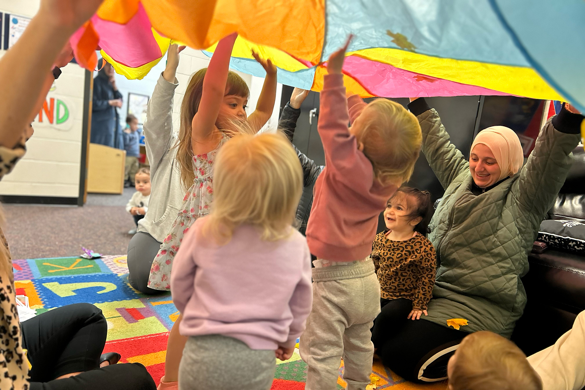 Young children in a classroom play under a parachute held by an adult