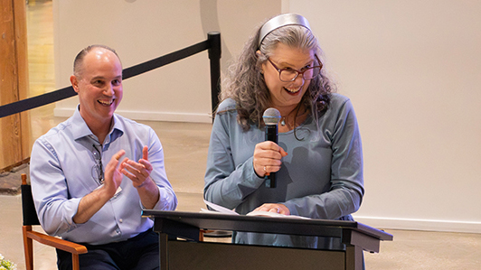 A woman standing to speak at a podium with a man clapping behind her