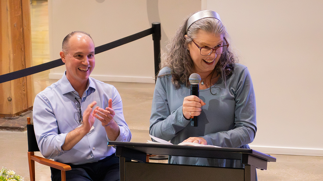 A woman standing to speak at a podium with a man clapping behind her