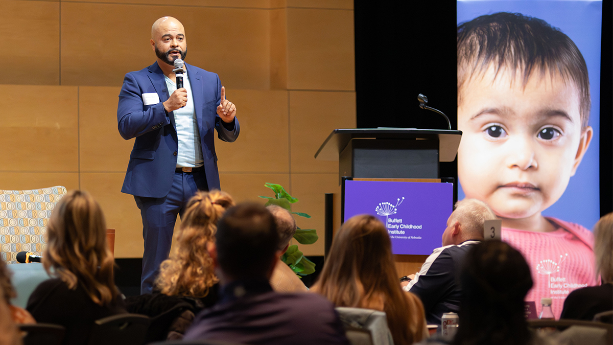 A man in a blue suit holds a microphone and speaks onstage at the 2026 Superintendents' Early Childhood Plan annual convening