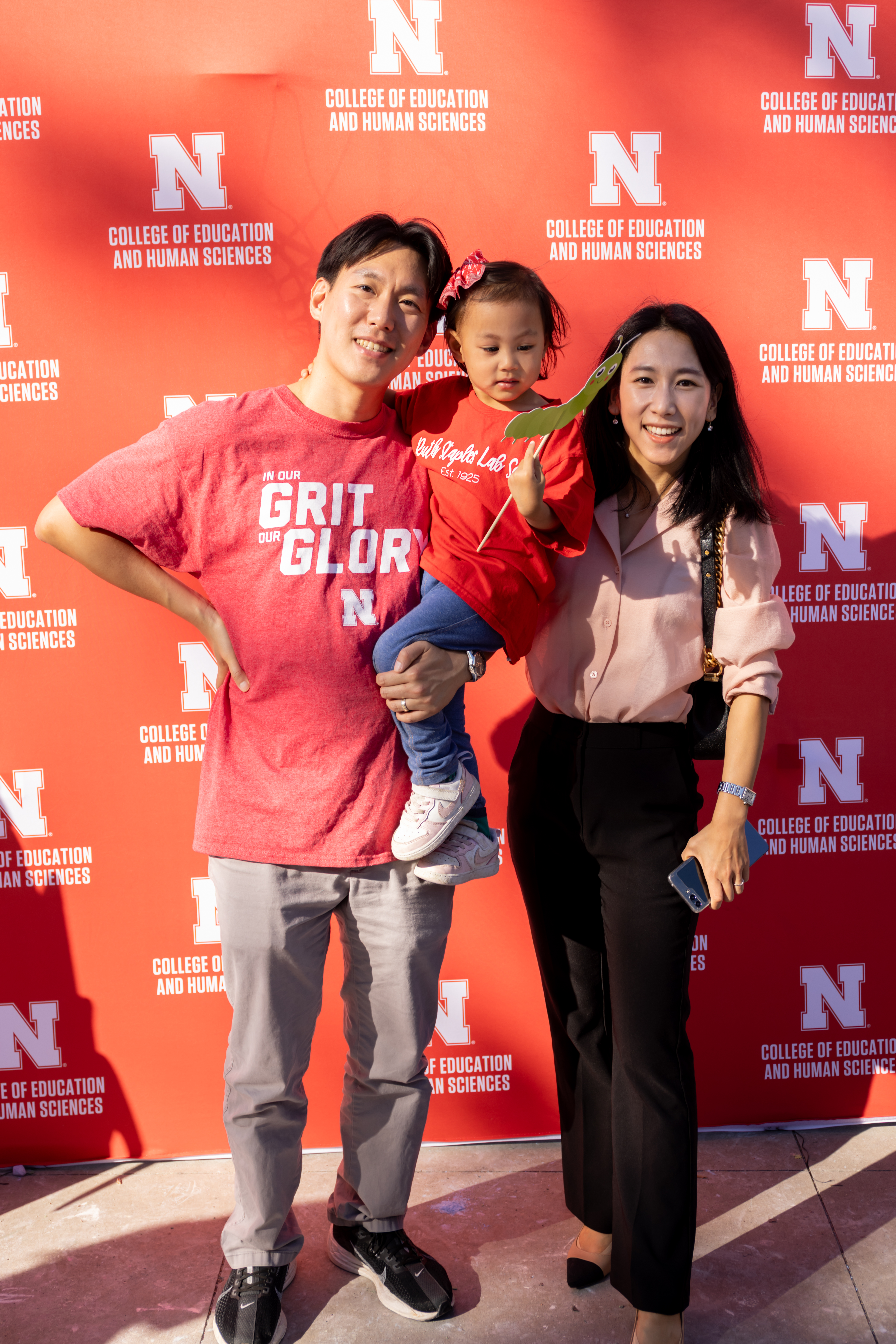 Two adults and a child pose in front of a red University of Nebraska backdrop