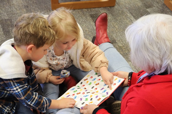 Two young children look at and touch a book being held by an early childhood educator 