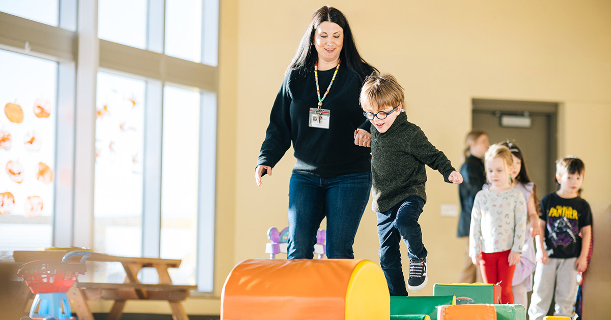 Preschool-aged children jump over play obstacles with the help of their teacher 