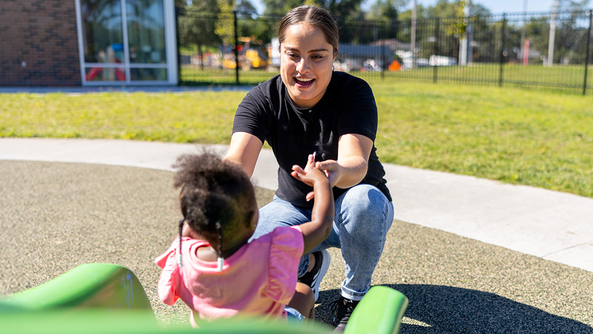 A female child care provider helps a young girl down a green slide outside