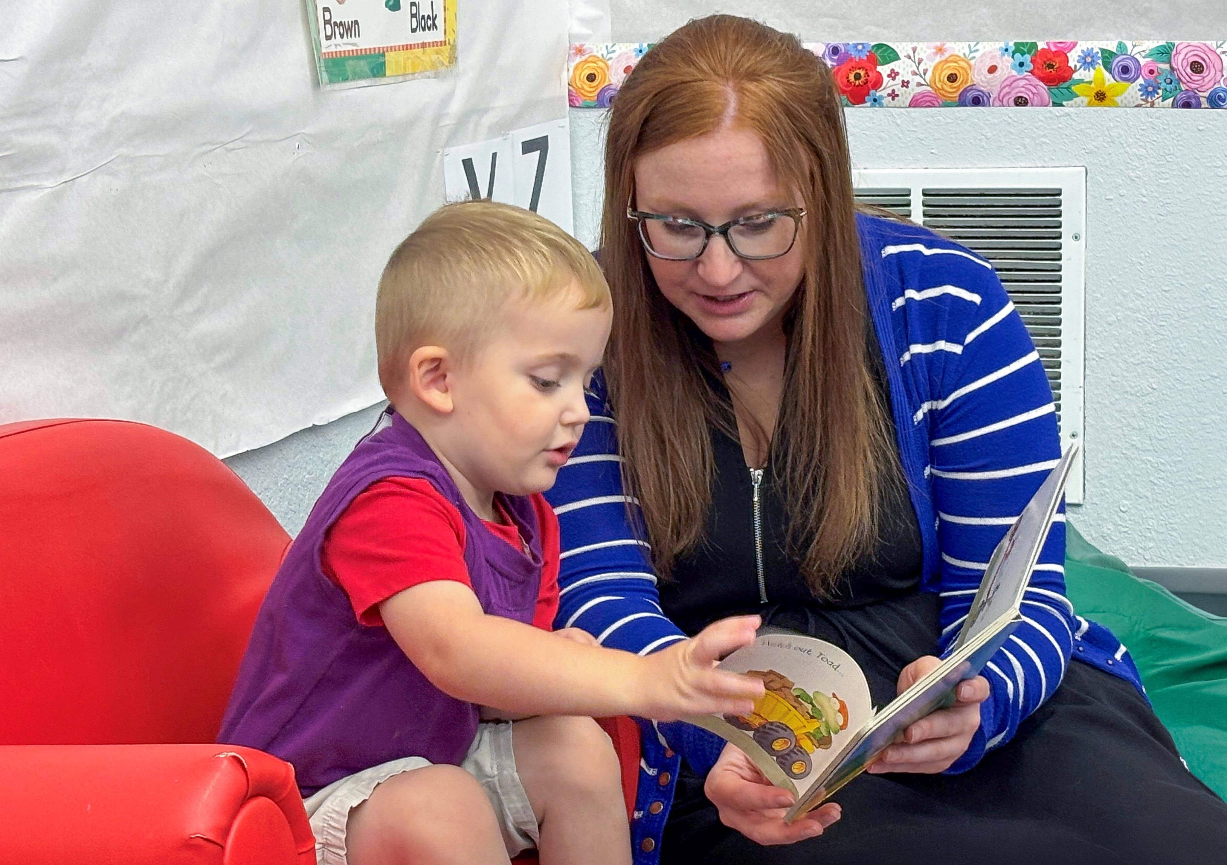Teacher reads a picture book with a young child, who points at an illustration while sitting together in a classroom reading area.