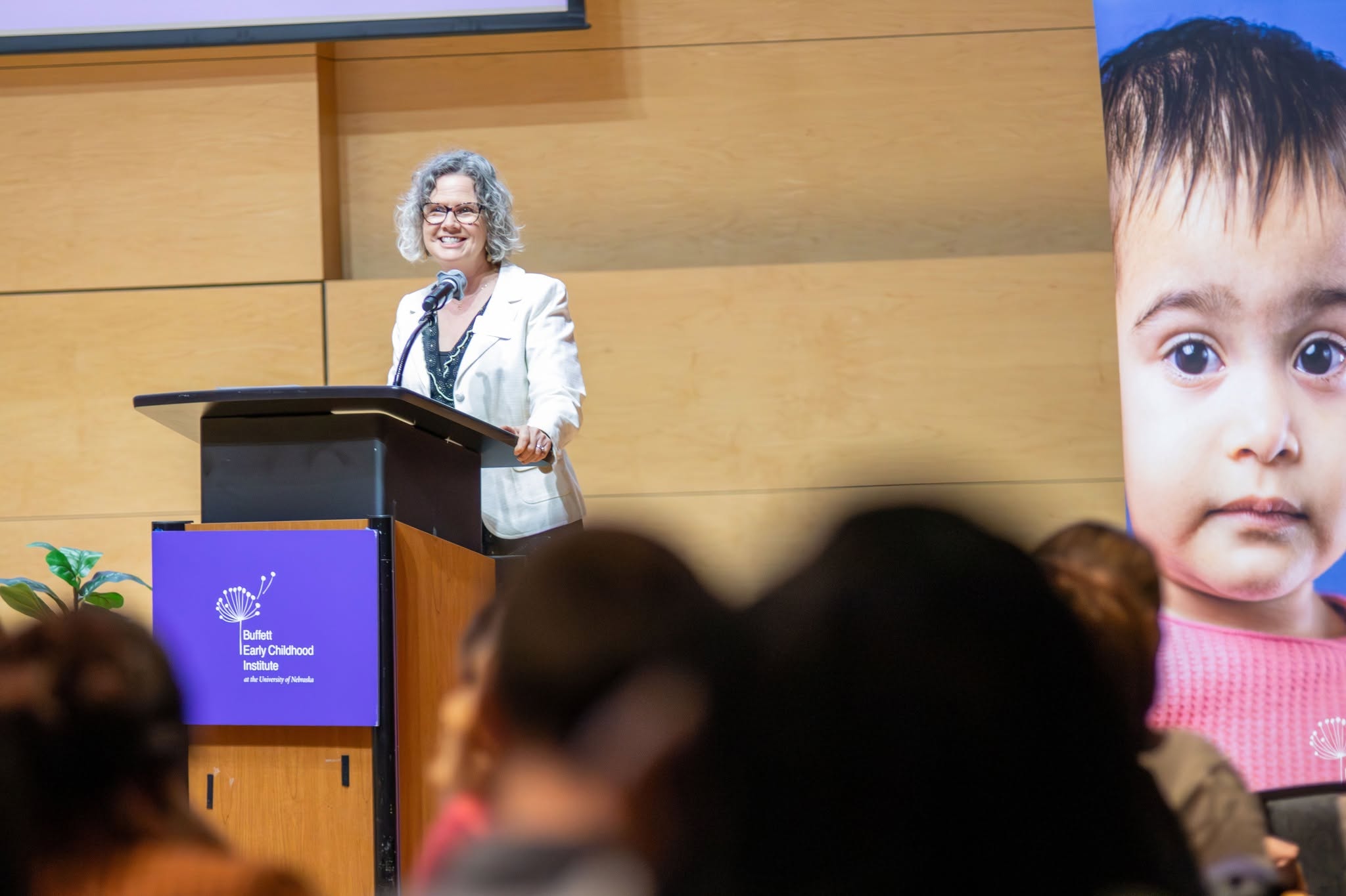 Speaker Amy Schmidtke stands at a podium with a microphone during a conference, with an audience in the foreground and a large banner showing a child’s face beside the stage.