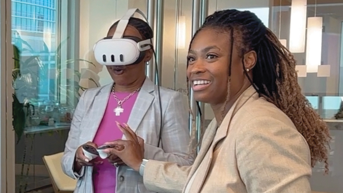 One woman wearing a VR headset and holding controllers while the other smiles and gestures nearby.