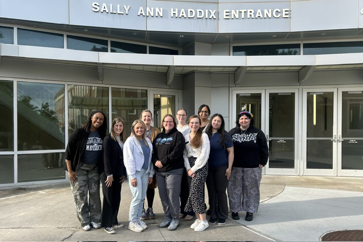 Group of students and program leaders from the University of Nebraska at Omaha posing outside a campus building entrance with the sign “SALLY ANN HADDIX ENTRANCE” above the doors.