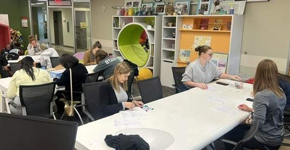 A group of people working and studying at tables in a modern room with shelves of supplies and a bright green circular reading nook.
