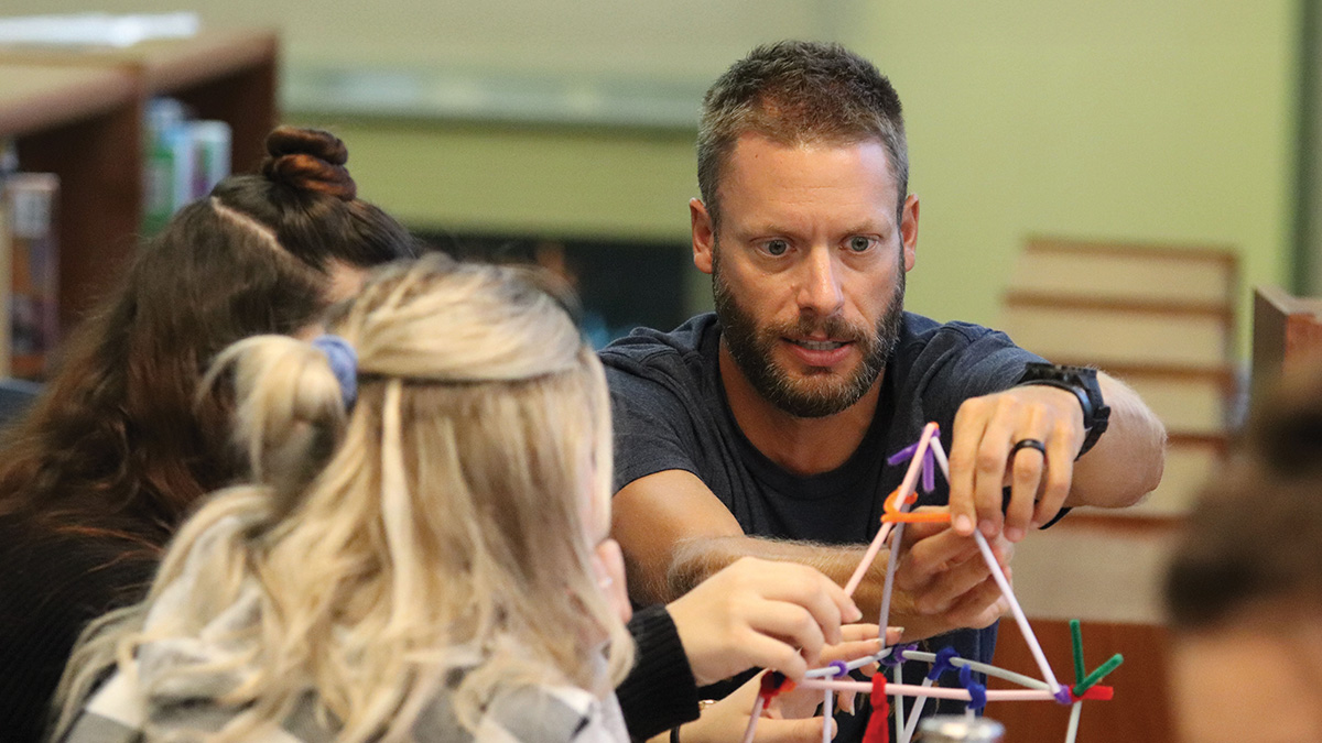 Teacher helps students build a geometric structure with colorful plastic rods and connectors in a classroom.