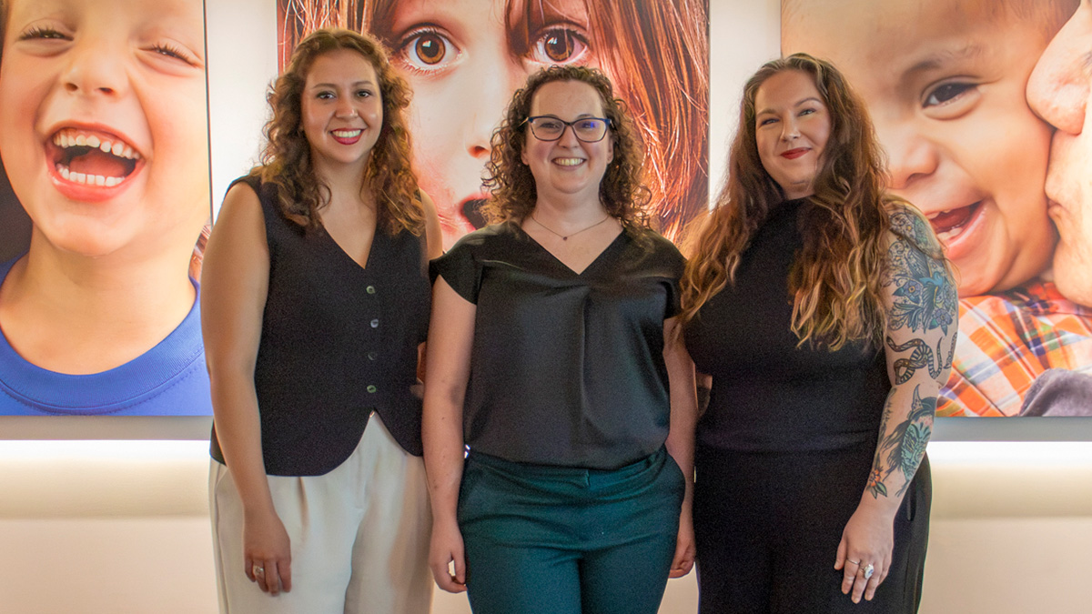 Three women standing and smiling in front of large wall photos of children’s faces.