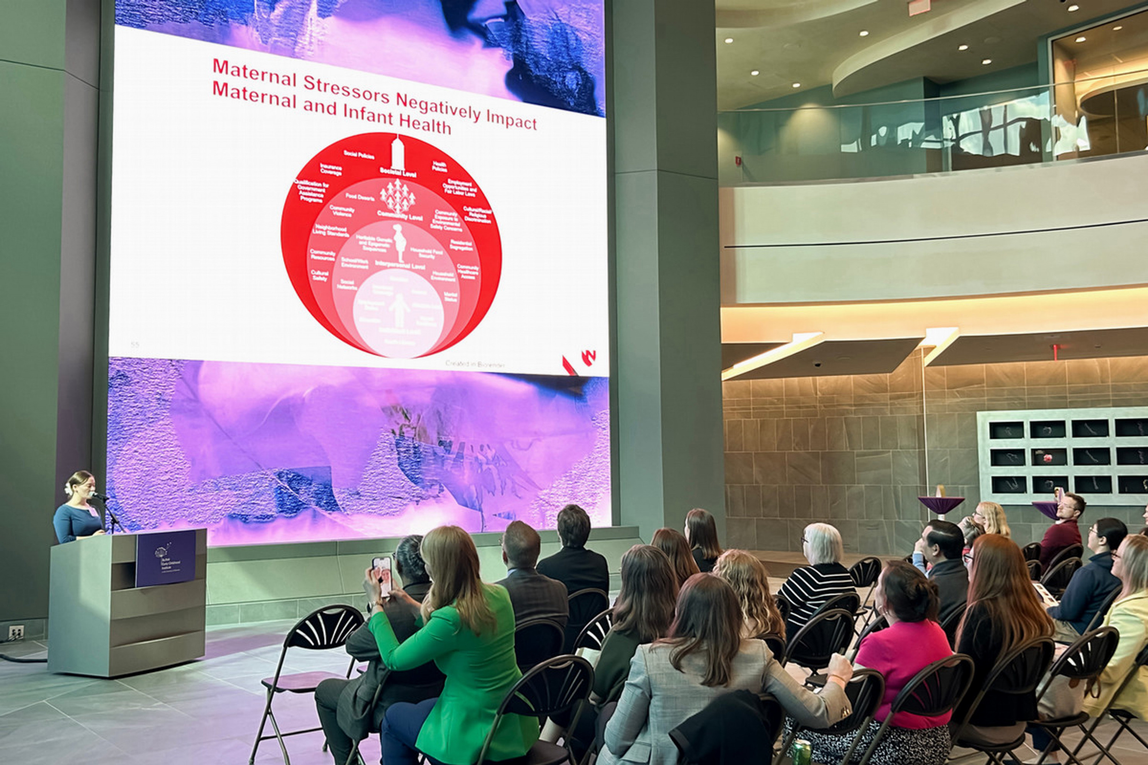 Speaker at a podium presents to a seated audience in a large atrium, with a projected slide about maternal stressors and maternal and infant health.