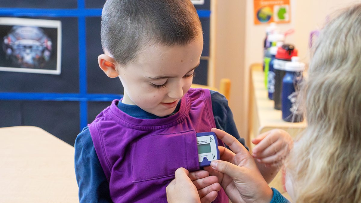 A preschool teacher helps a young child attach a small electronic device to the front of a purple vest in a classroom setting.