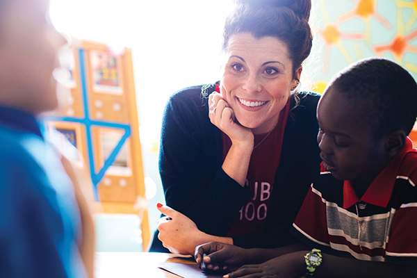 Teacher leans in and smiles while helping a student with schoolwork at a classroom table.