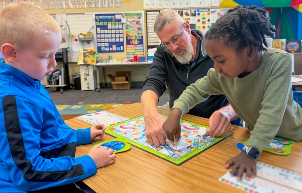 Teacher helps two students play a board game together at a classroom table.