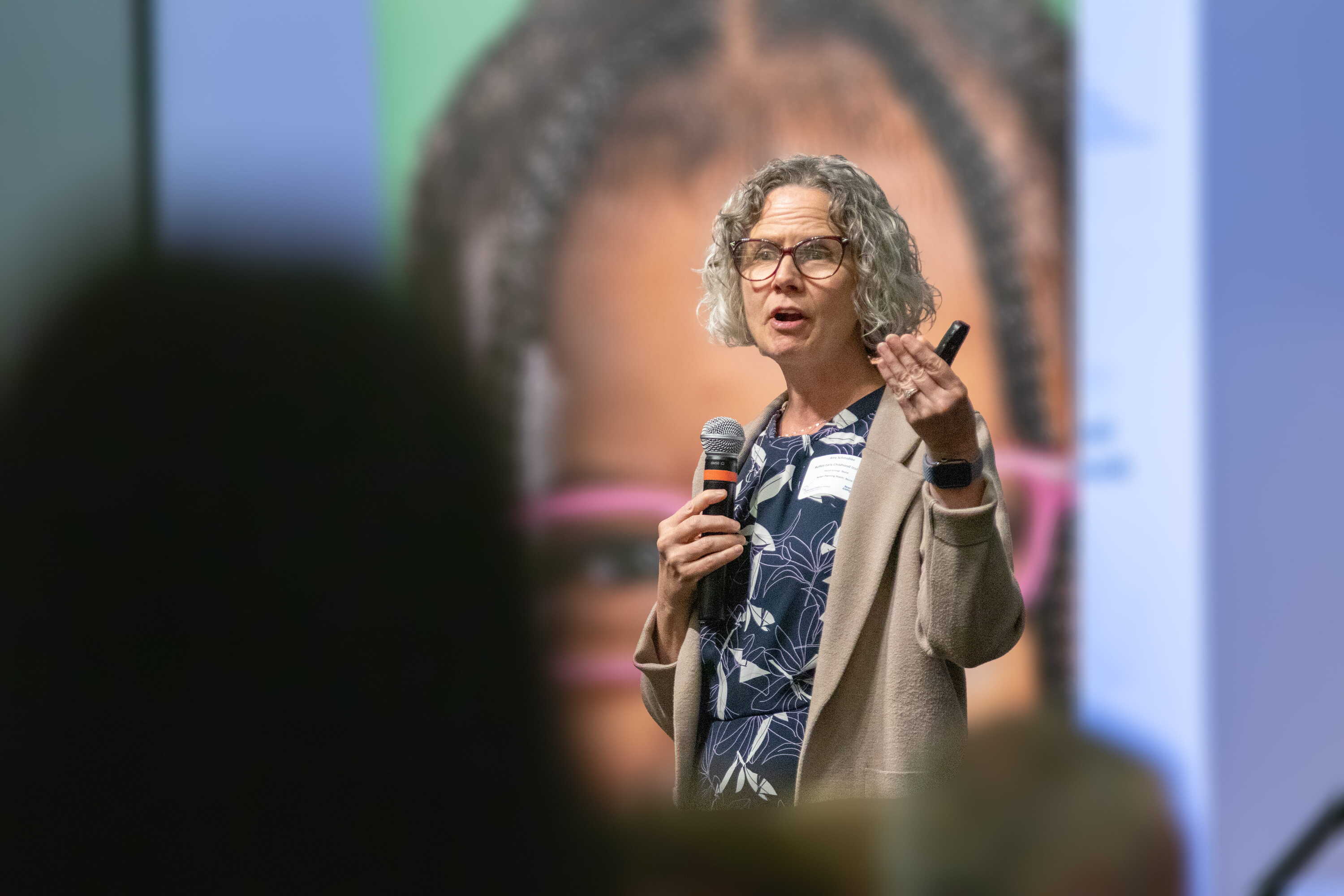 Amy Schmidtke on stage holding a microphone and gesturing while giving a presentation, with a blurred audience in the foreground.
