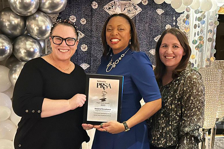 Three women pose at an awards event in front of a silver-and-white balloon backdrop, holding a Nebraska PRSA plaque.