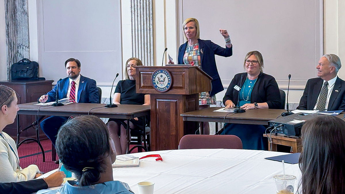 Panel discussion in the U.S. Senate, with Raleigh Smith Duttweiler speaking at a podium bearing the U.S. Senate seal while several panelists sit at tables and an audience listens.