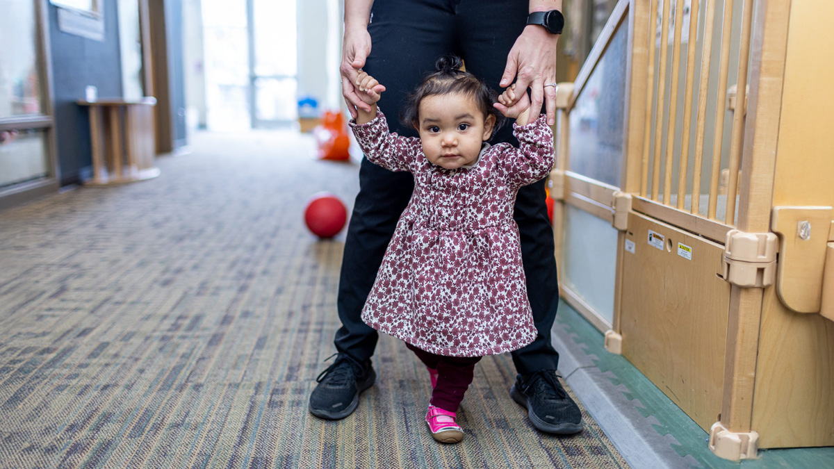 A child care provider helps a baby walk forward