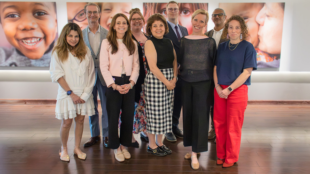 Group of nine professionals posing indoors in front of large wall photos of children’s faces.