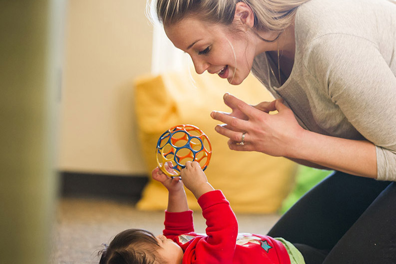 Woman kneels and smiles while playing with a baby holding a colorful lattice ball on the floor.