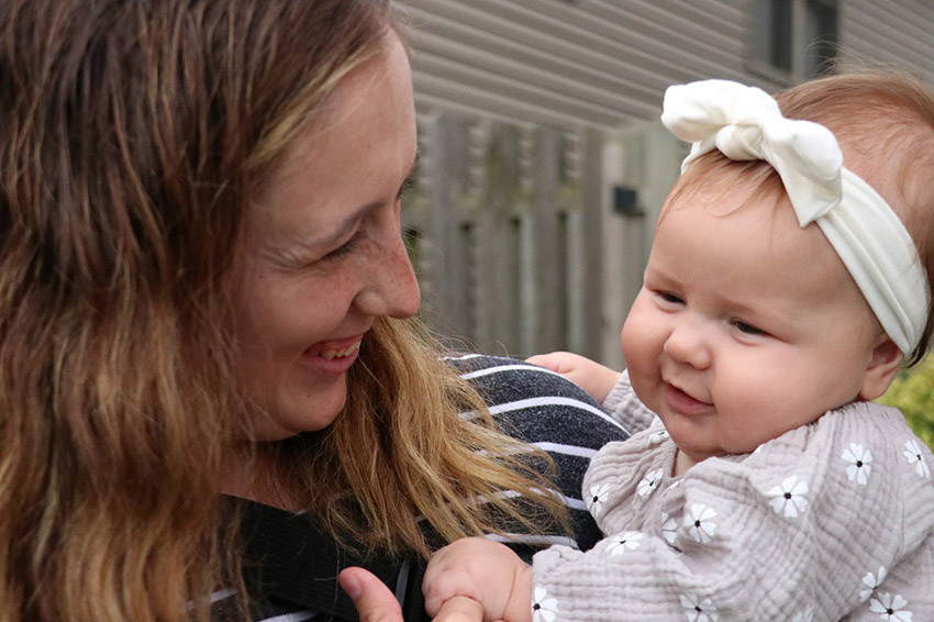 Woman smiling while holding a baby wearing a white headband bow outdoors.