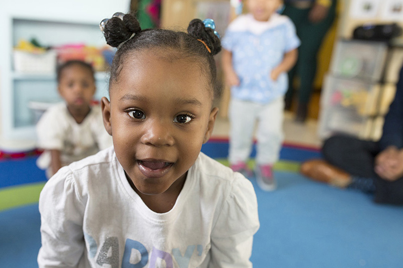 Toddler with hair in two buns smiles toward the camera while other children play in a daycare classroom.