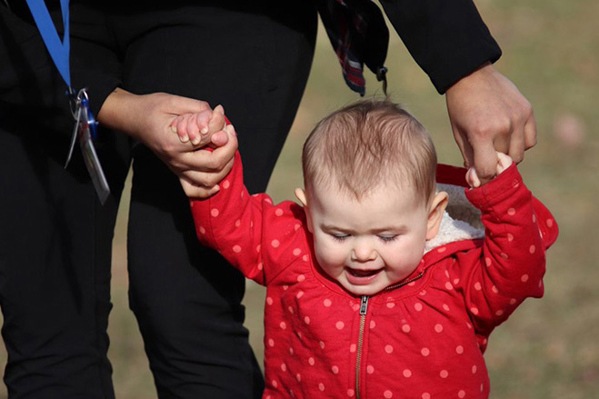 Adult holding a baby’s hands as the child walks outdoors, wearing a red polka-dot jacket.