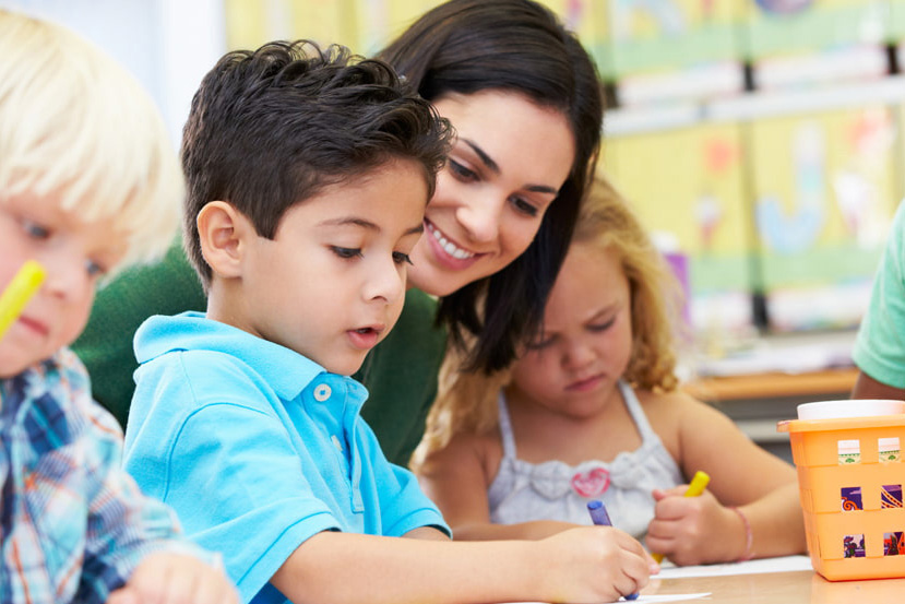 Teacher leaning in to help young children as they draw and write at a classroom table.