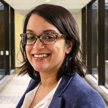 Smiling person wearing glasses and a blue blazer in a bright office hallway.