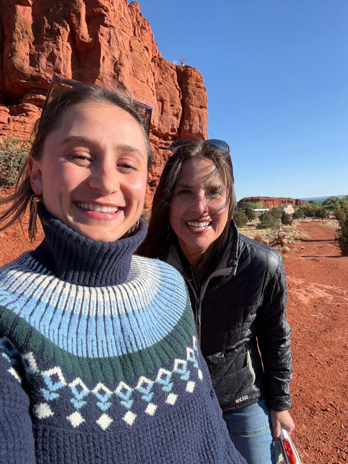 Alexandra Gregory, left, and Michelle Sarche, right, smile for a selfie on a sunny desert trail with red rock cliffs in the background.