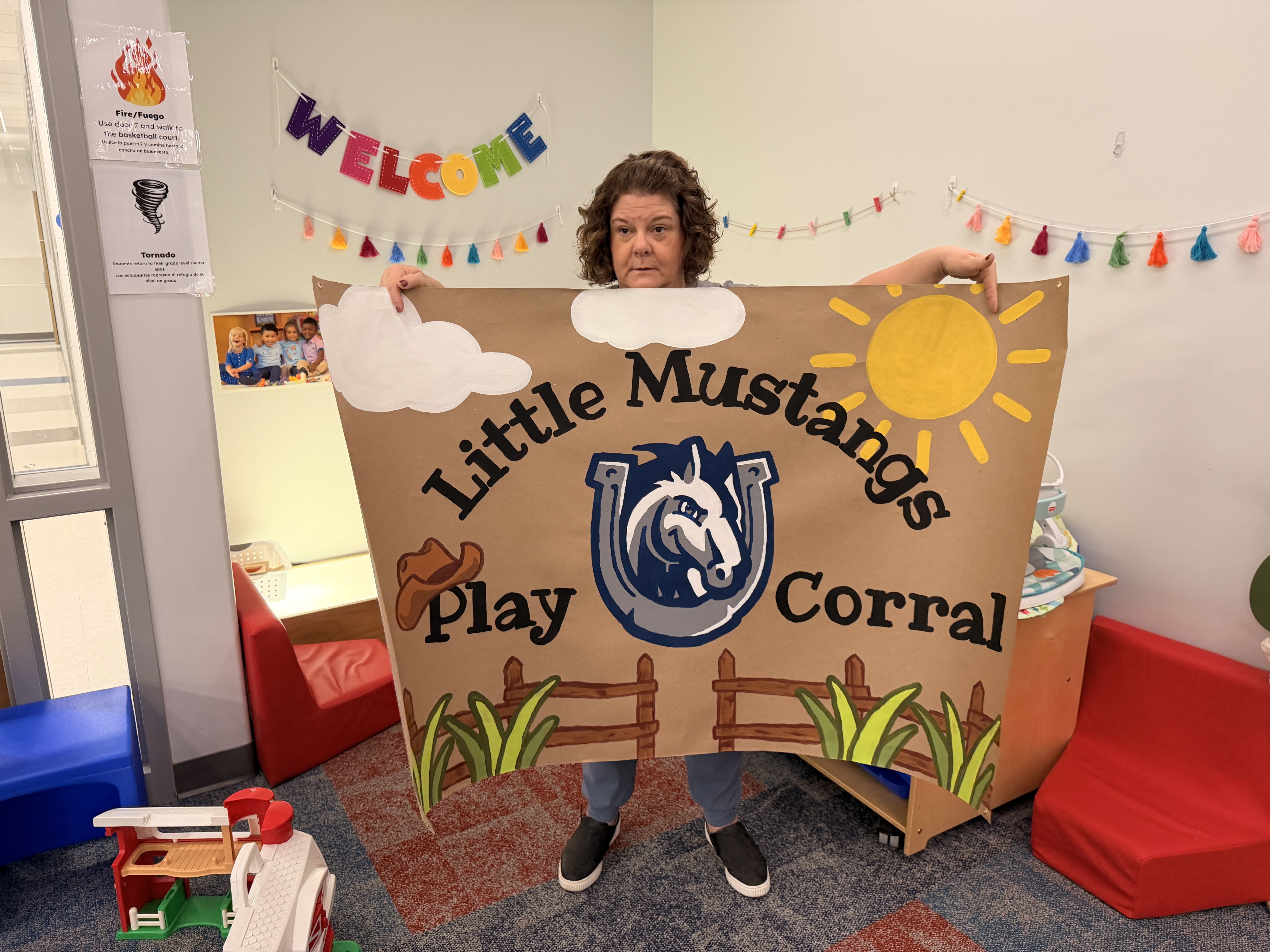 An early childhood educator holds a large sign that says "Little Mustangs Play Corral"