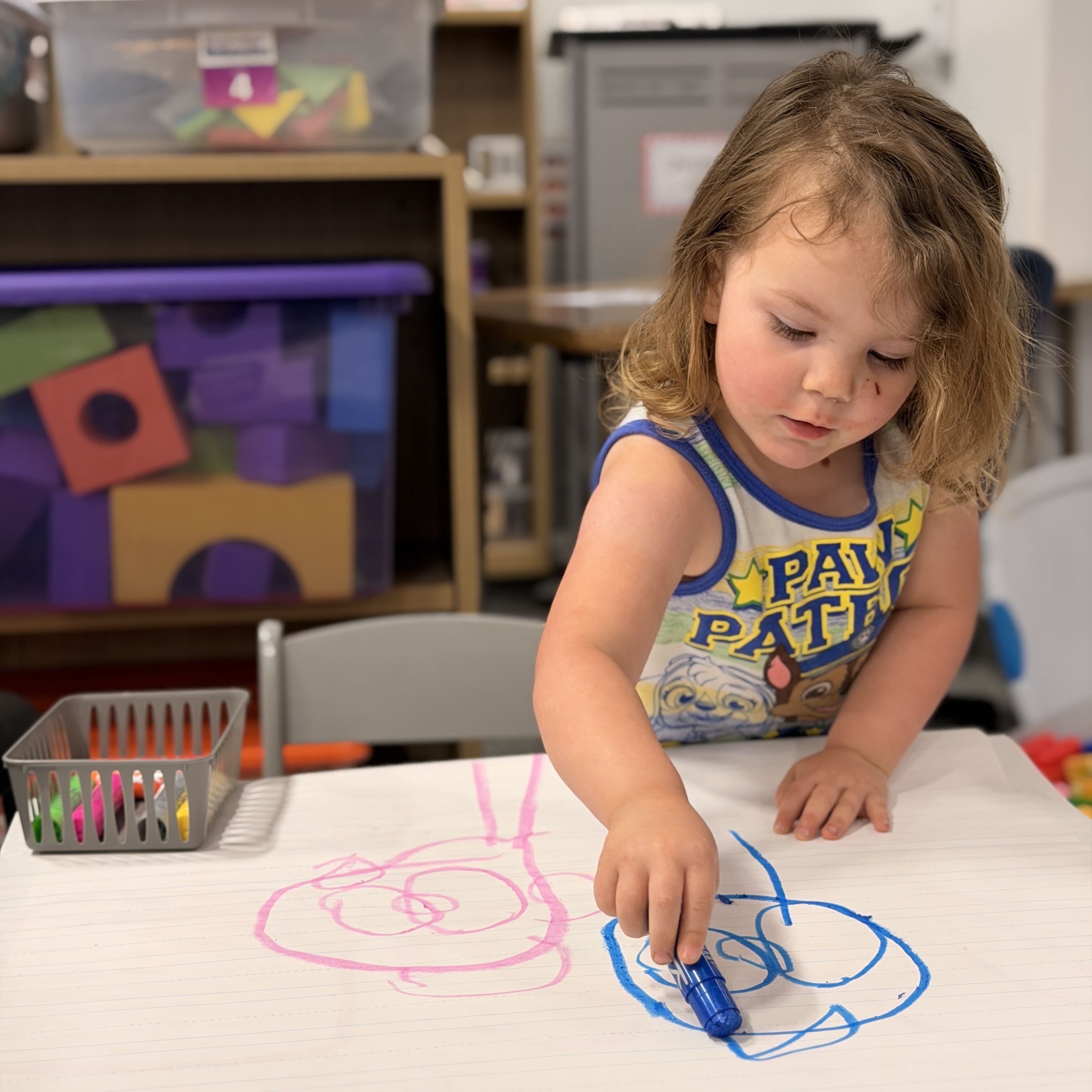 A toddler-age girl draws with crayons on a large, white sheet of paper