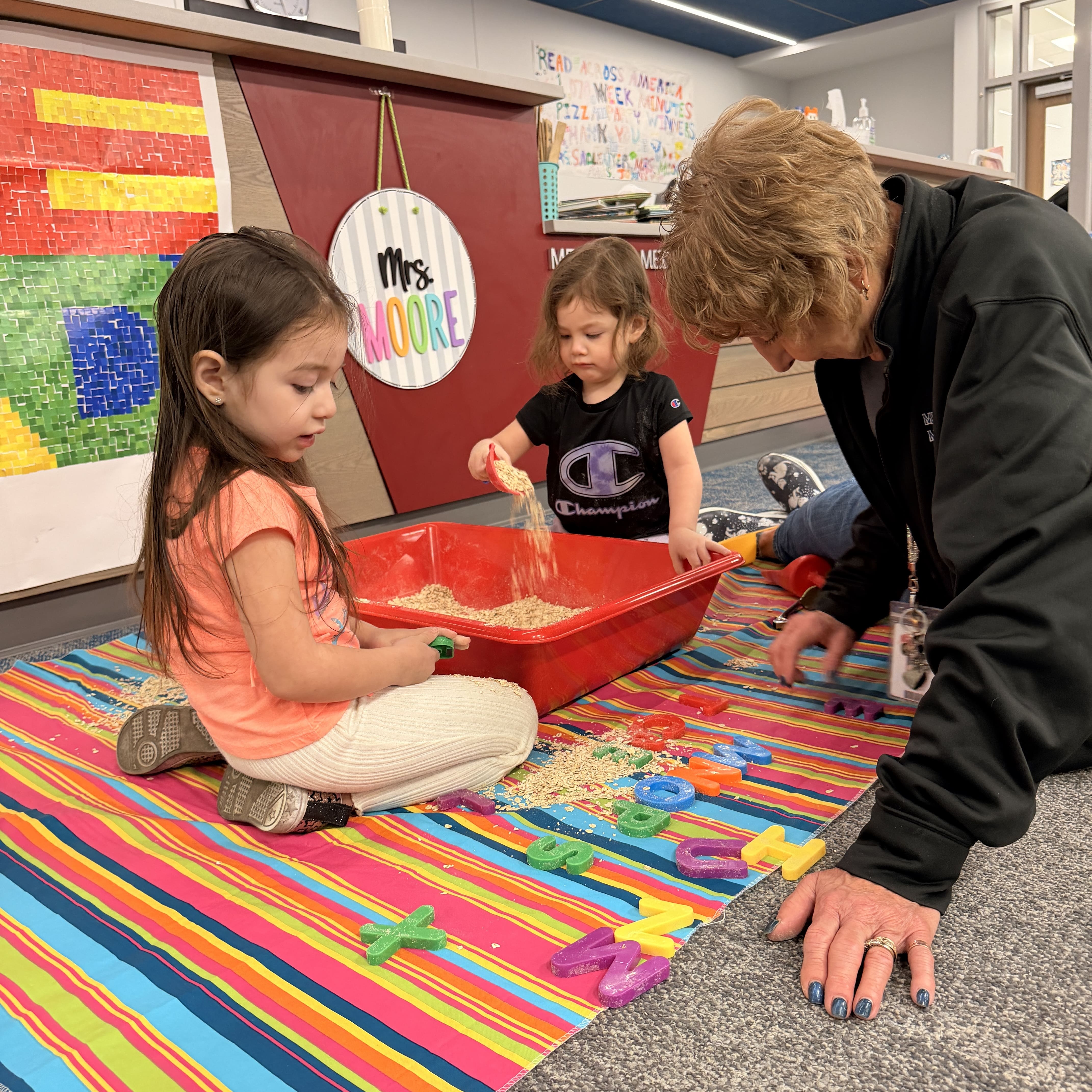 Two young children and an adult volunteer play with toys in a red container filled with sand on the ground