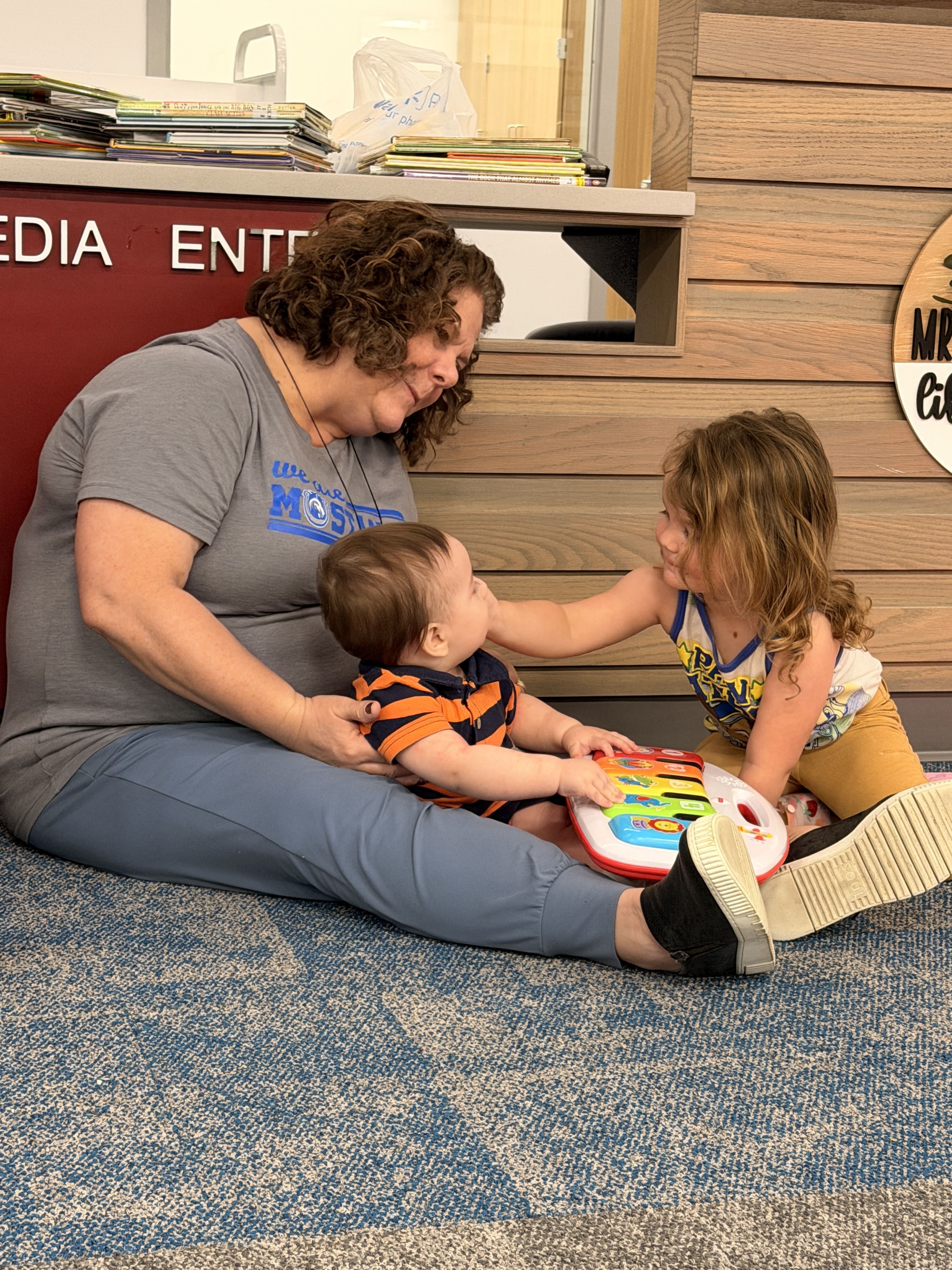 An early educator holds a baby in front of her while a toddler interacts with the baby