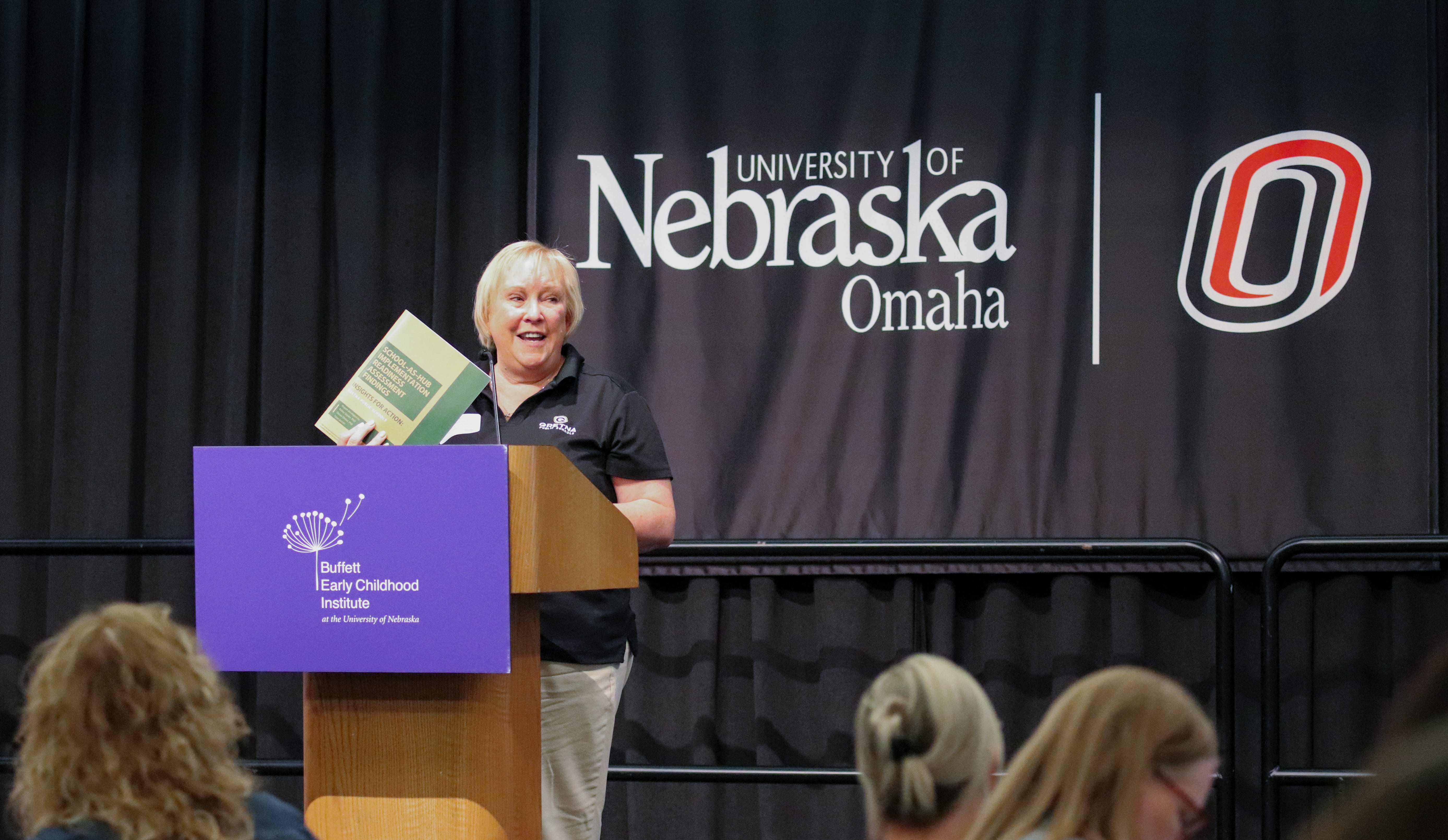 Speaker at a podium holds up a book during the 2026 Superintendents' Early Childhood Plan collaboration summit event at the University of Nebraska at Omaha.