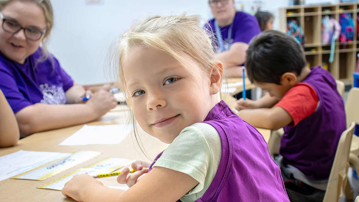 Young student in a purple vest looks toward the camera while working at a classroom table, with other children and a teacher in the background.