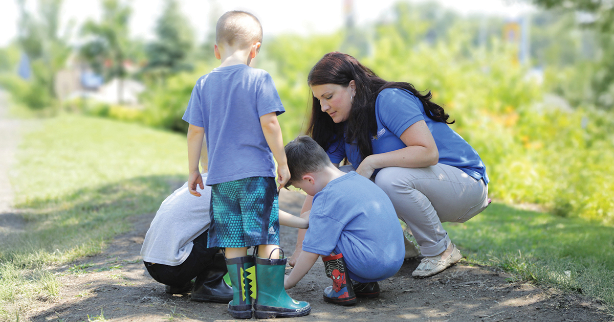 Woman crouches outdoors with three children as they examine something on the ground along a grassy path.
