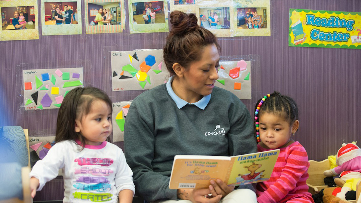 Teacher reads a picture book with two young children seated on the floor in a classroom reading corner.