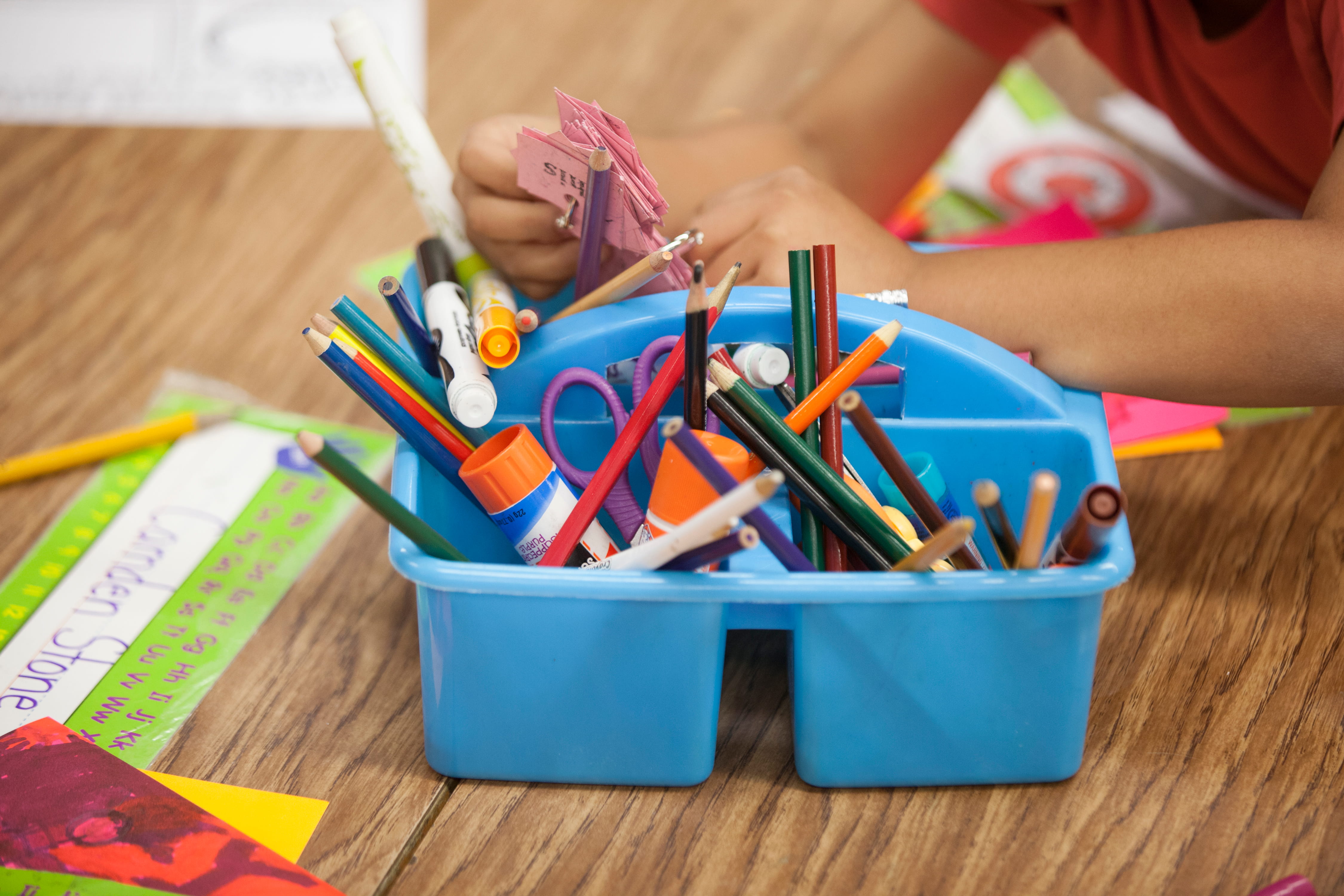 Blue plastic caddy filled with pencils, markers, scissors, and glue sticks on a classroom table.