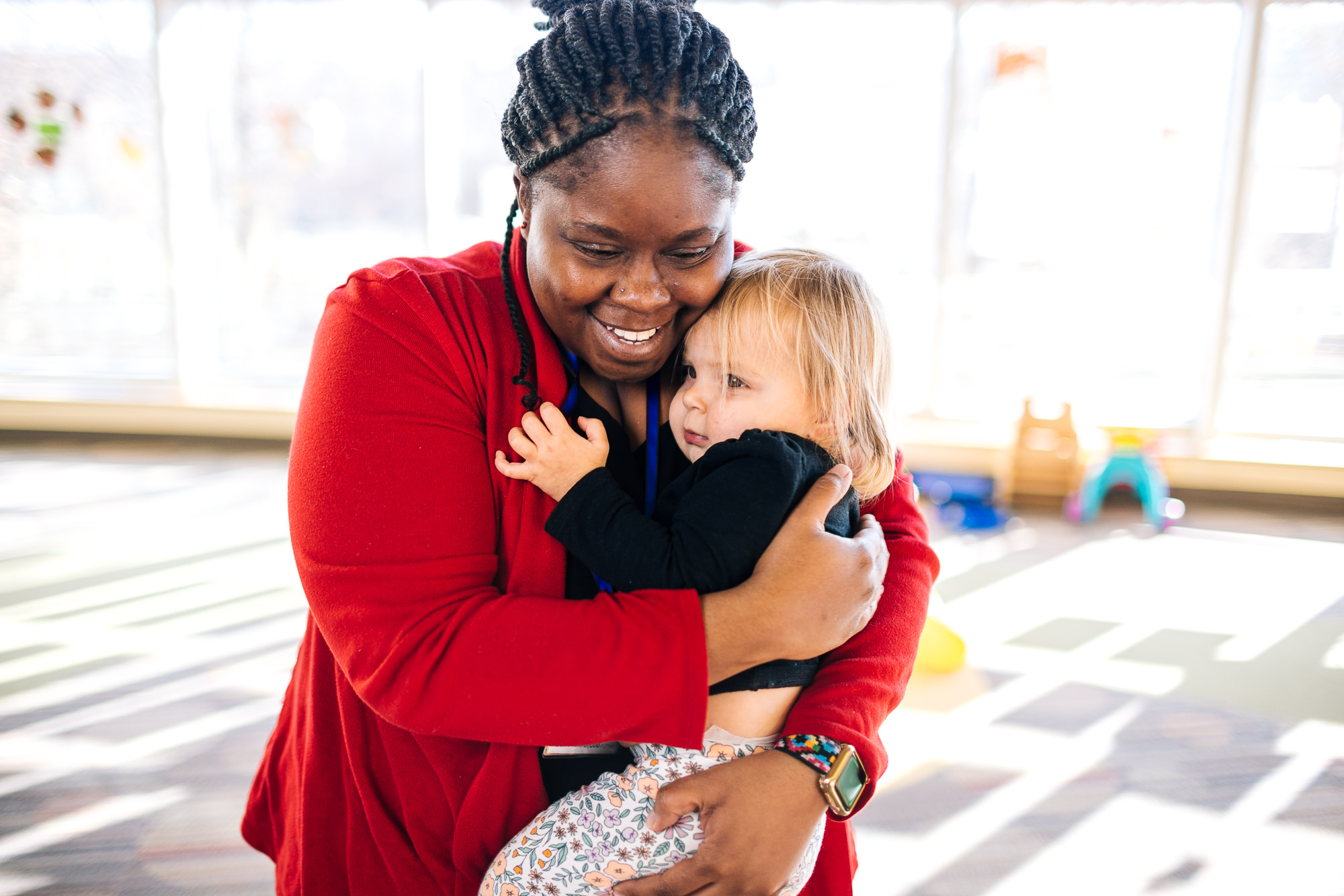 Caregiver hugs a young child in a bright daycare classroom.