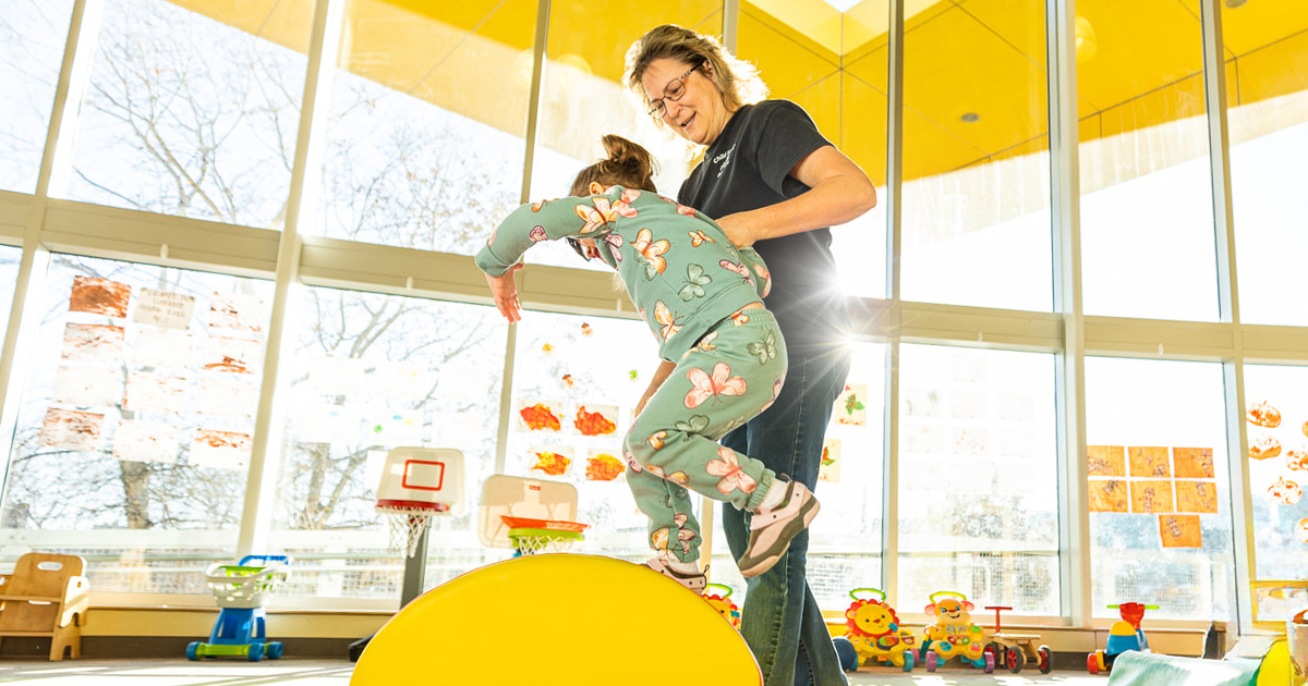 A child care provider helping a child jump over a play obstacle