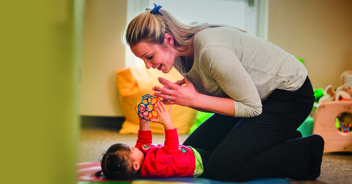 Woman playing with a toddler lying on a mat and holding a colorful ball toy.